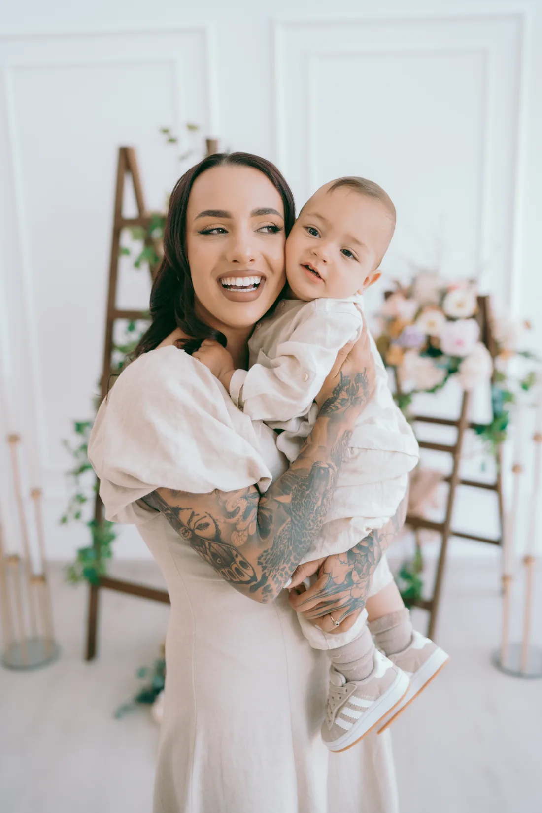 Naomi Crawford smiling while holding her son Theodoros, both wearing pale linen.